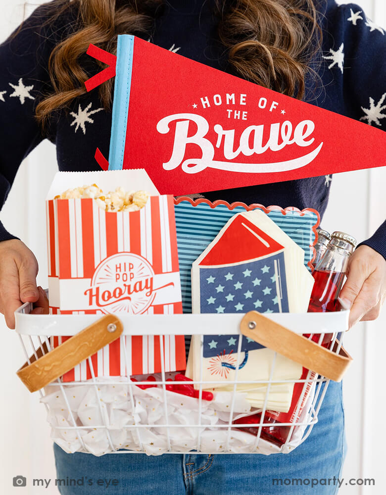 A lady holding an occasion bin filled with festive patriotic themed party goods including Momo Party's 15" Home of the Brave red felt pennant by My Mind's Eye, blue stripe scallop-edged plates, rocket shaped guest napkins, red striped popcorn treat boxes with "hip hip hooray" on it, these are perfect decorations for a Memorial Day celebration or 4th of July Independence Day party!