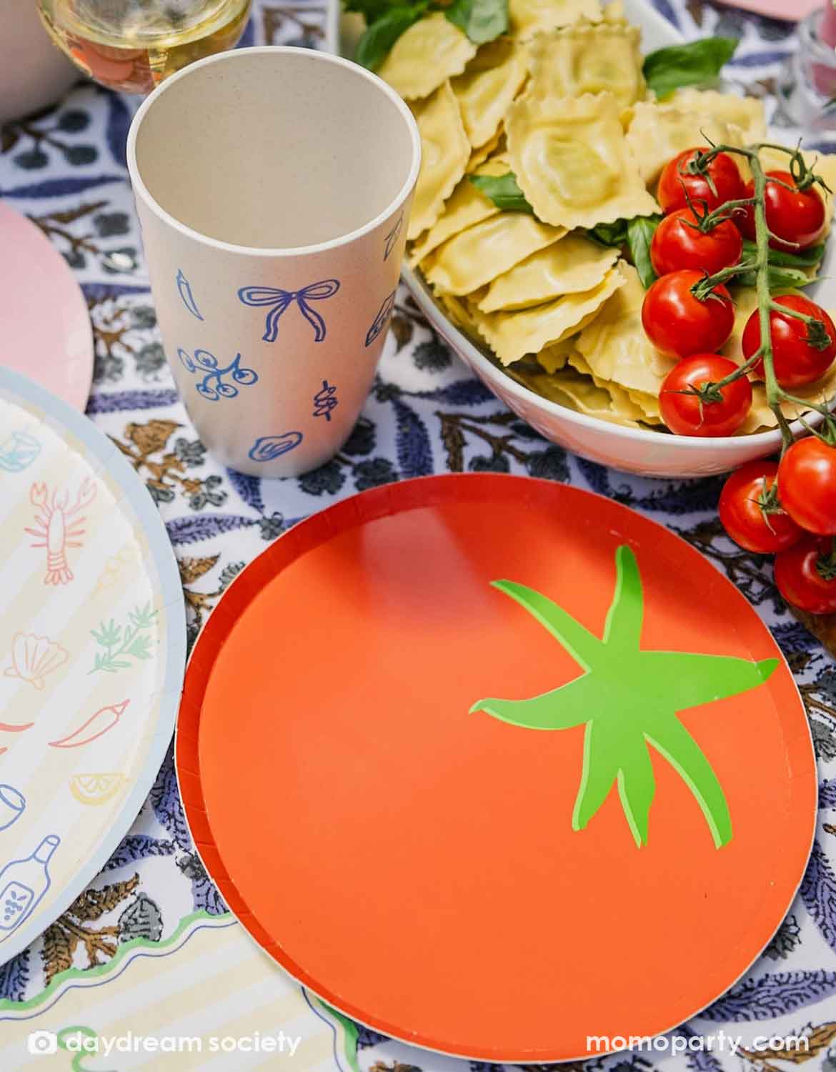 A festive Italian-inspired party table setting with Momo Party's 8" tomato shaped plate surrounded by a bowl of pastas and tomatoes with plates, cups and drinks around.