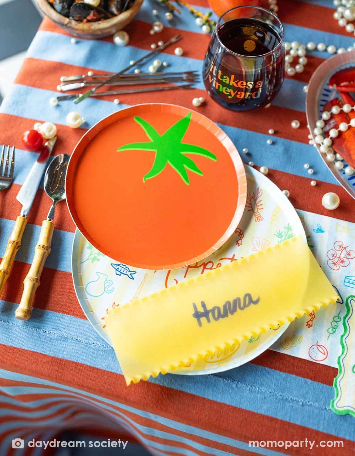 A festive Italian-inspired party table setting with Momo Party's 8" tomato shaped plate surrounded by a bowl of pastas and tomatoes with plates, cups and drinks around on a blue and red striped tablecloth.