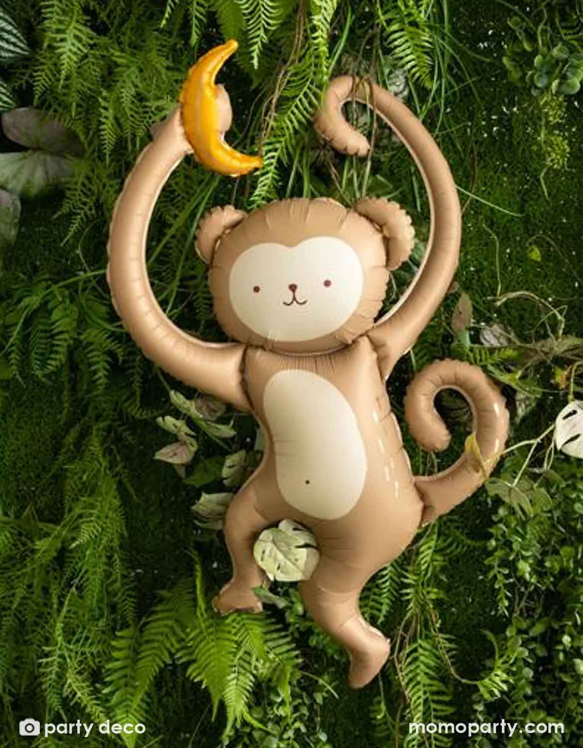 Momo Party's 35" monkey with a banana shaped Foil balloon by Party Deco hanging from a branch with green leaves against a green leafy jungle background.