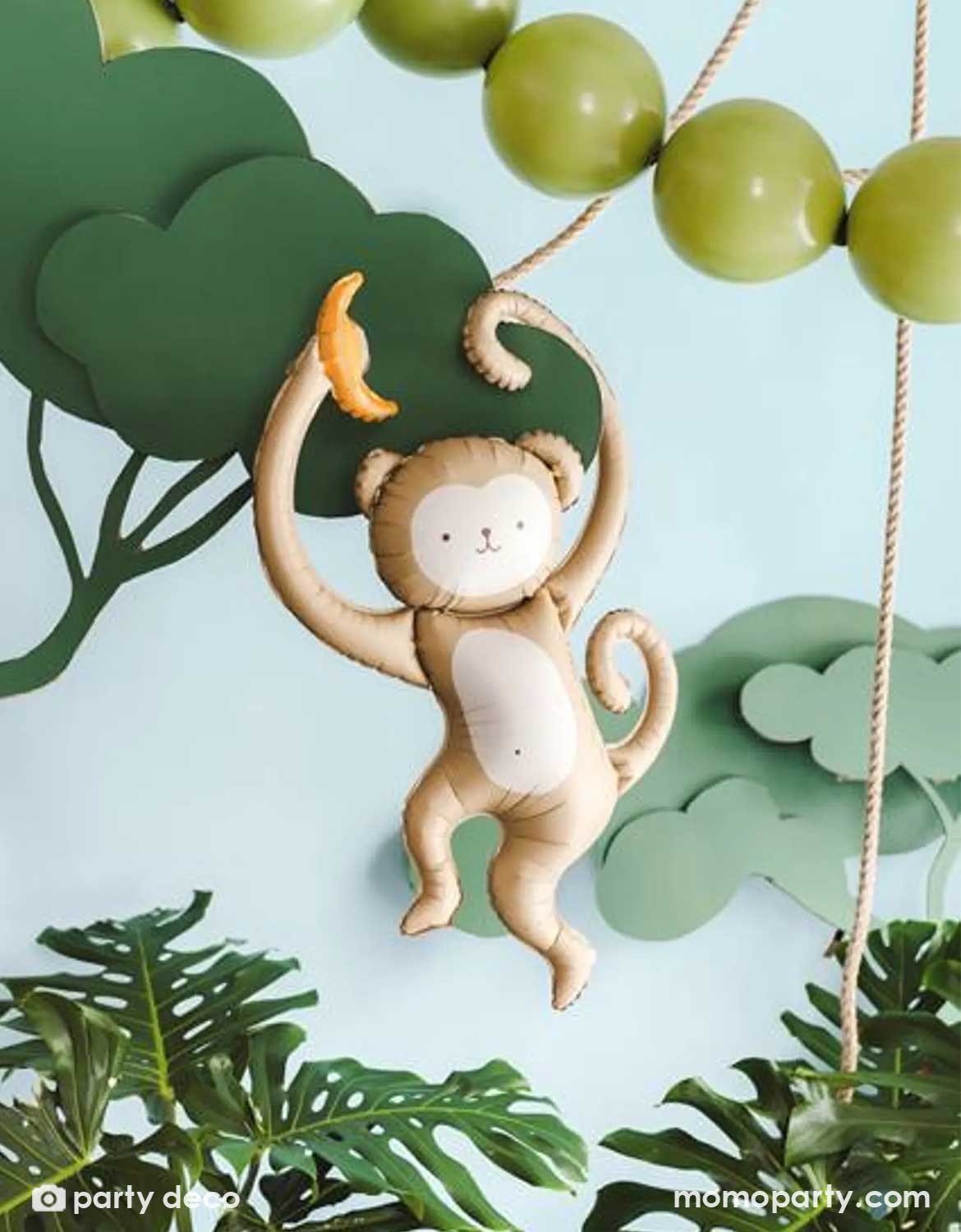 Momo Party's 35" monkey shaped Foil balloon with a banana hanging from a branch with green leaves and green latex balloon string on a paper decorative jungle background.