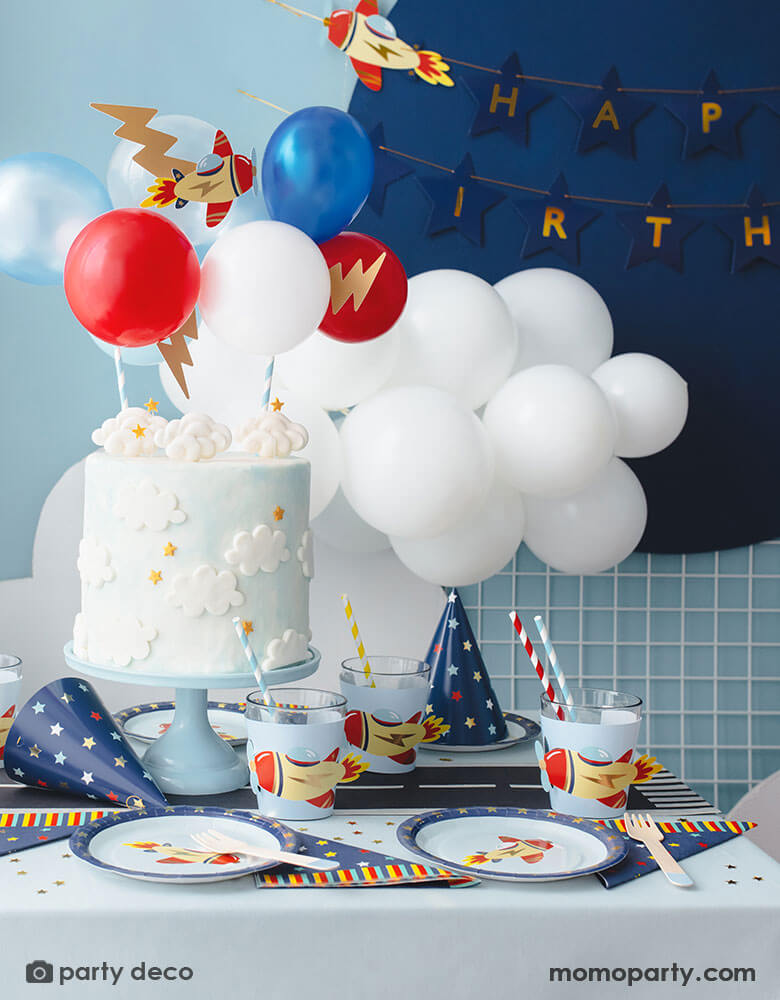 A kid's airplane birthday party decoration featuring Momo Party's plane themed party supplies and decorations by Party Deco, including 7" vintage airplane round paper plates, navy and star around large napkins and party hats, vintage airplane cup sleeves wrapped around party cups with red and blue striped paper straws. In the middle of the table is a birthday cake topped with airplane toppers and colorful mini balloons. In the back there's a blue backdrop with happy birthday banner hung on it. 