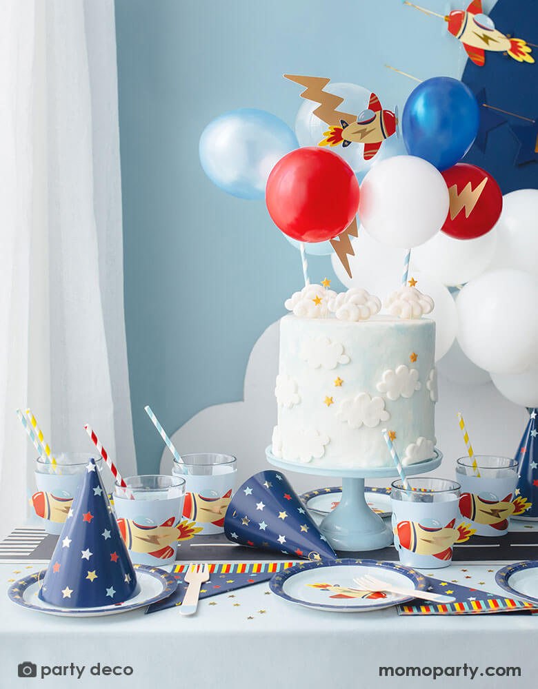 A kid's airplane birthday party decoration featuring Momo Party's plane themed party supplies and decorations by Party Deco, including 7" vintage airplane round paper plates, navy and star around large napkins and party hats, vintage airplane cup sleeves wrapped around party cups with red and blue striped paper straws. In the middle of the table is a birthday cake with cloud designs topped with airplane toppers and colorful mini balloons. Makes this an adorable inspo for kid's aviation birthday party.