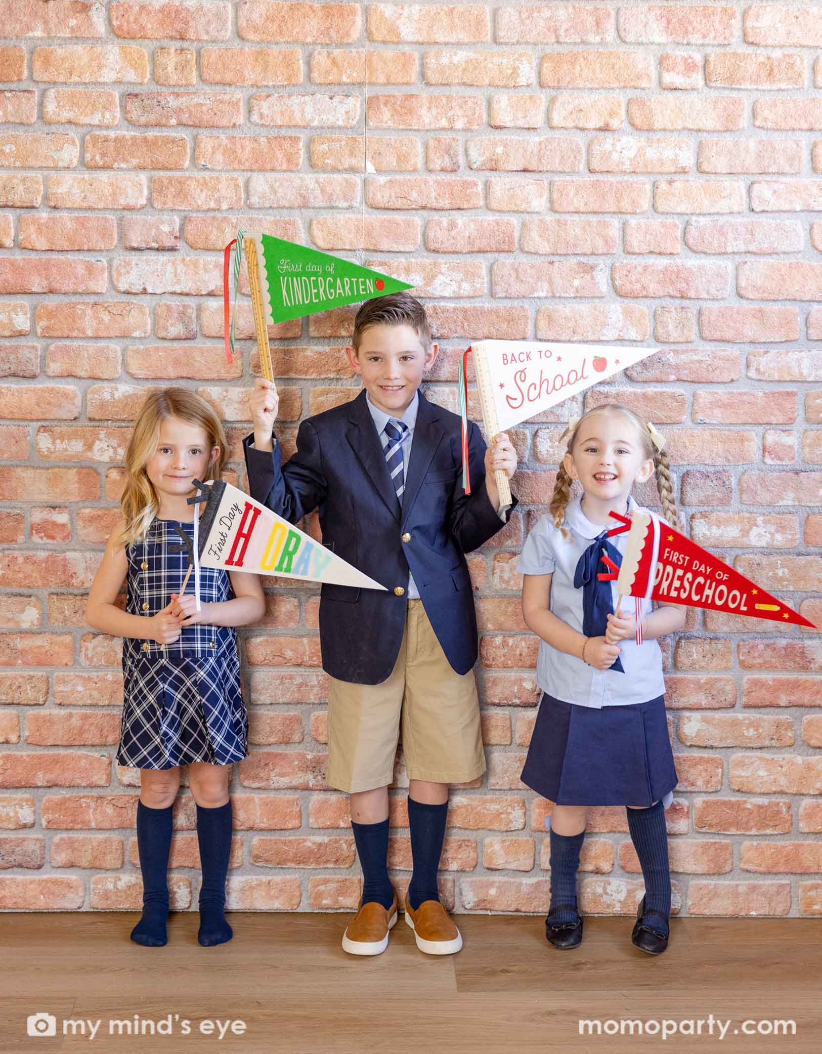 Three children in their uniforms holding school-themed party pennants including the green first day of kindergarten party pennant, the ruler back to school party pennant, the red first day of preschool party pennant and the white first day hooray pennant. They're smiling and standing against a brick wall ready for the new school year!