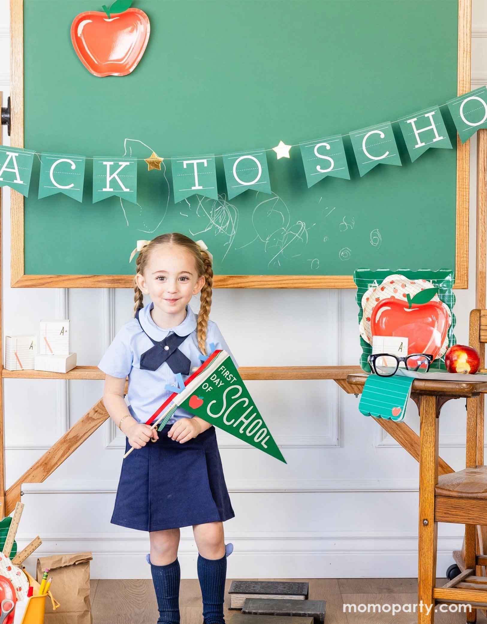 A school-aged girl in her blue uniform holding Momo Party's green first day of school apple felt pennant standing in a classroom setting decorated with a bunch of school themed party supplies from Momo Party including apple shaped plates, alphabet dinner plates and matching napkins. Behind her is a classroom chalkboard with back to school party banner hung across with an apple shaped plate - an adorable and cute set up to celebrate kid's back to school season!