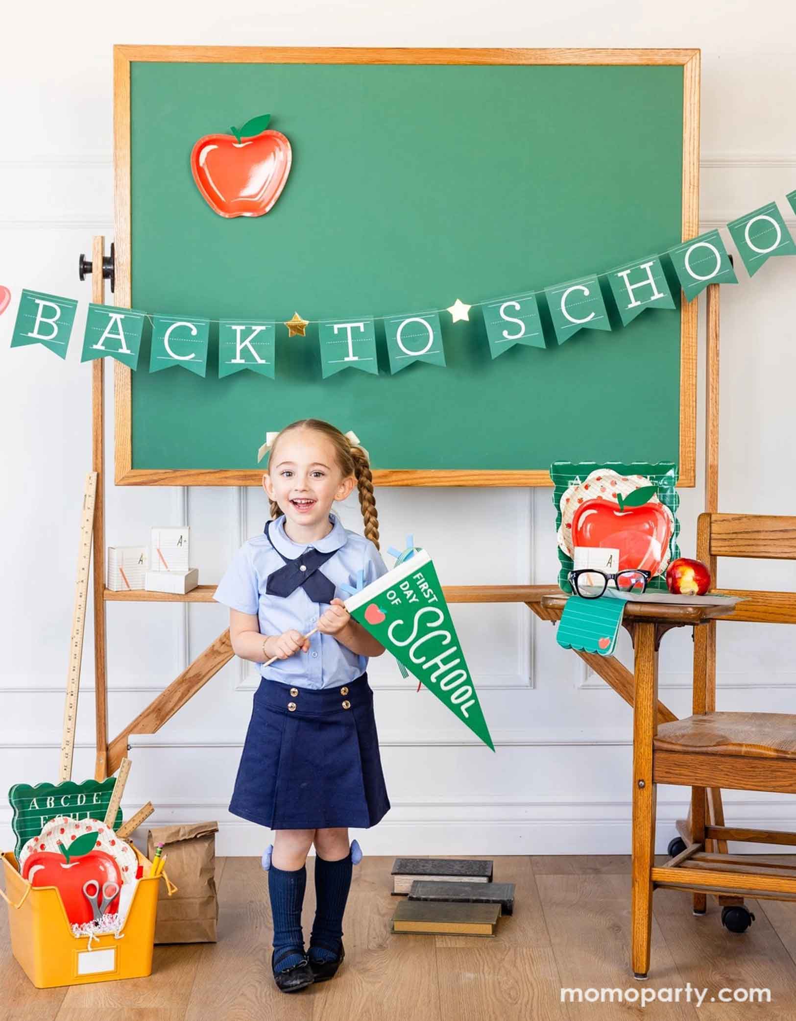 A school-aged girl in her blue uniform holding Momo Party's green first day of school apple felt pennant standing in a classroom setting decorated with a bunch of school themed party supplies from Momo Party including apple shaped plates, alphabet dinner plates and matching napkins. Behind her is a classroom chalkboard with back to school party banner hung across with an apple shaped plate - an adorable and cute set up to celebrate kid's back to school season!