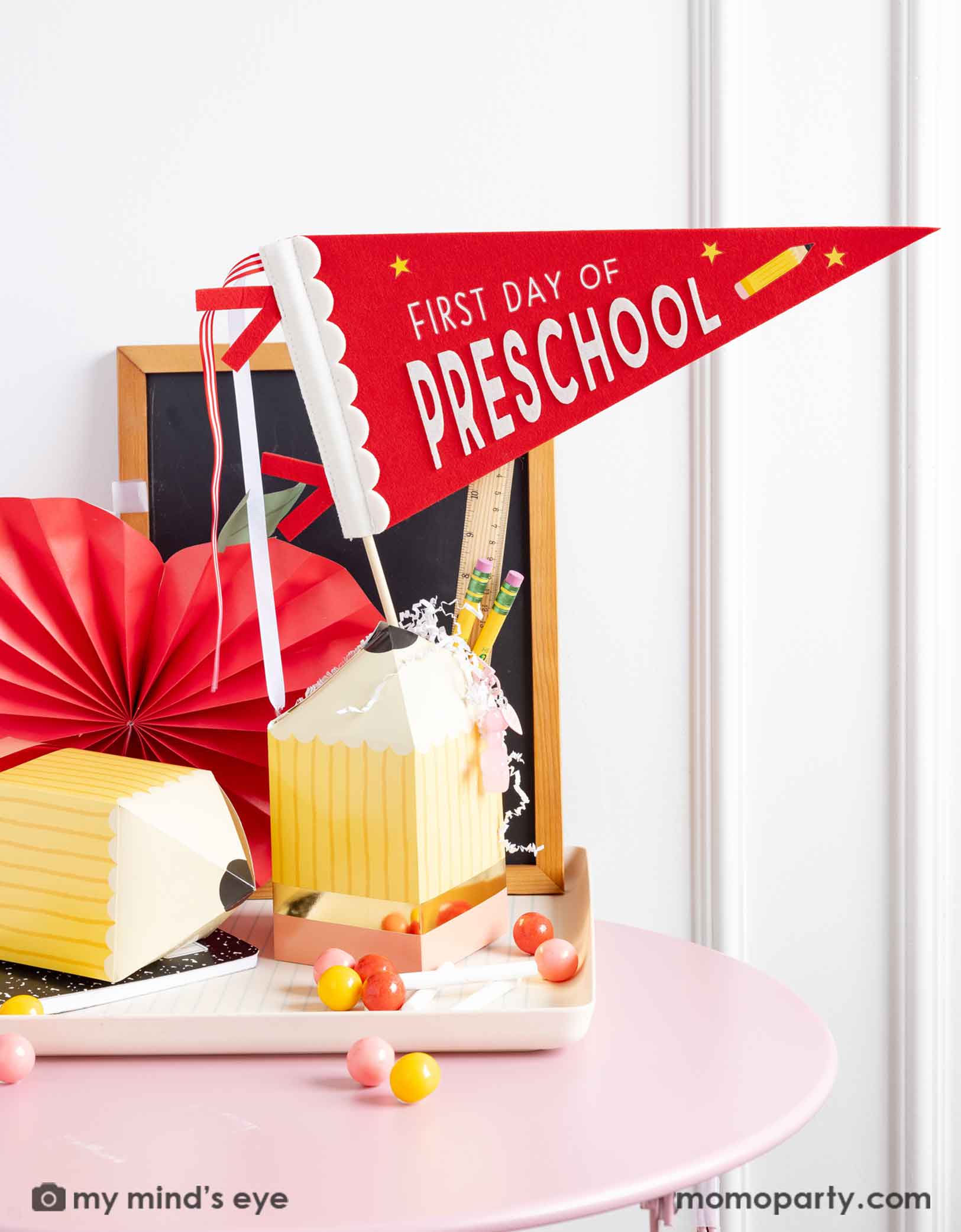 A festive back to school table set up decorated with various school themed party supplies from Momo Party including the red first day of preschool party pennant, pencil shaped treat boxes, notebook shaped bamboo party tray and apple shaped party fan. With a mini chalkboard in the back and some school supplies like pencils, rulers and some gumballs on the table, it creates a cute and modern look for a first day of school celebration.