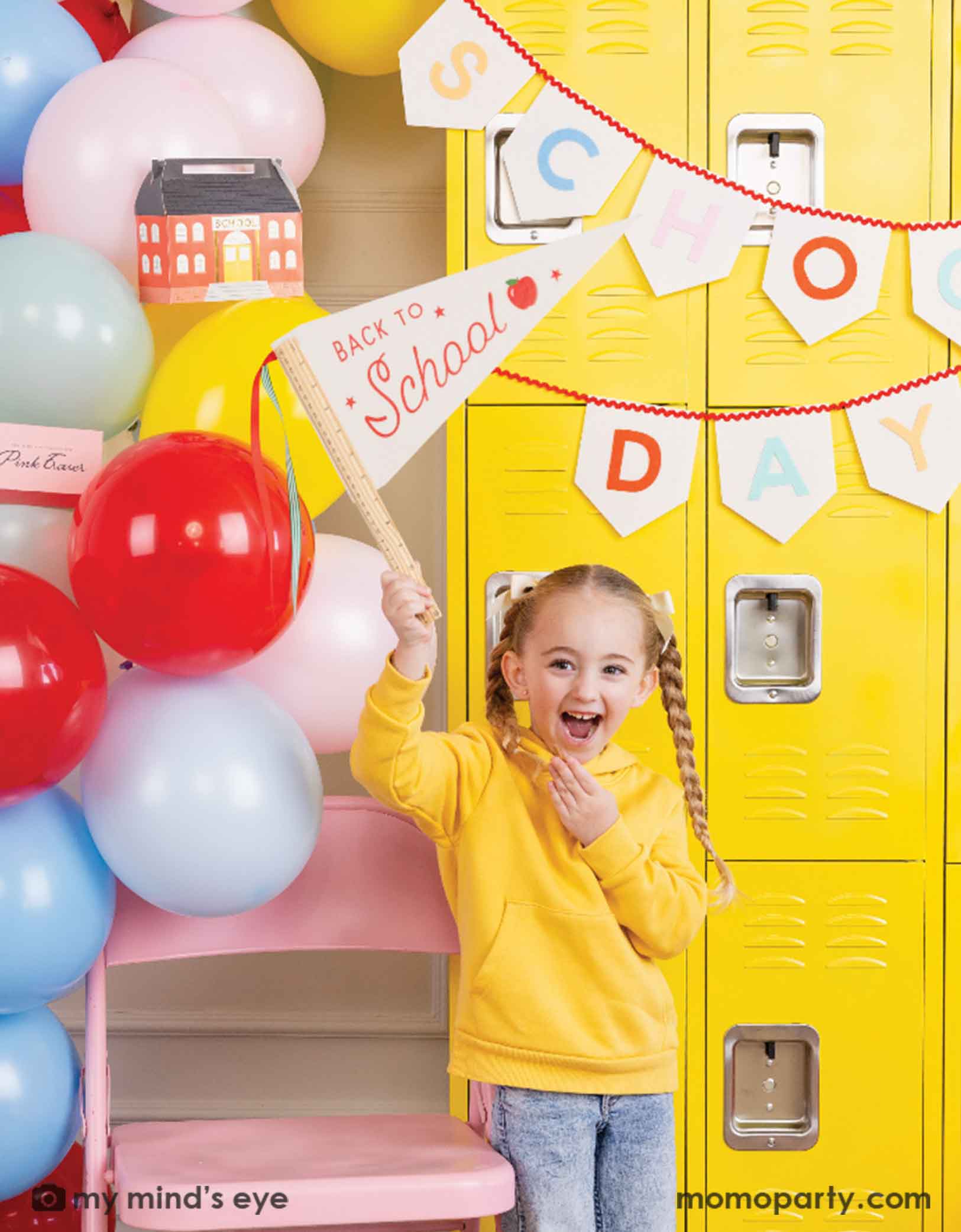 A girl in yellow shirt standing in front of a first day of school celebration decoration set up featuring various school themed party decors from Momo Party including School Days felt party banner and a festive balloon garland. She's holding the ruler back to school felt party pennant smiling happily. There's a yellow locker wall behind her and a pink folding chair. Making this a cute party decoration set up for kid's first day of school celebration.
