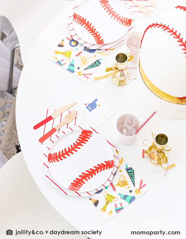 A sports themed party tablescape featuring Momo Party's 7.5 inch baseball plate layered on top of red striped plates,  napkins and sports themed napkins by Daydream Society. In the middle of the table there's a baseball shaped cake which is surrounded by mini trophies, all for a fun sports champion celebration for kids!