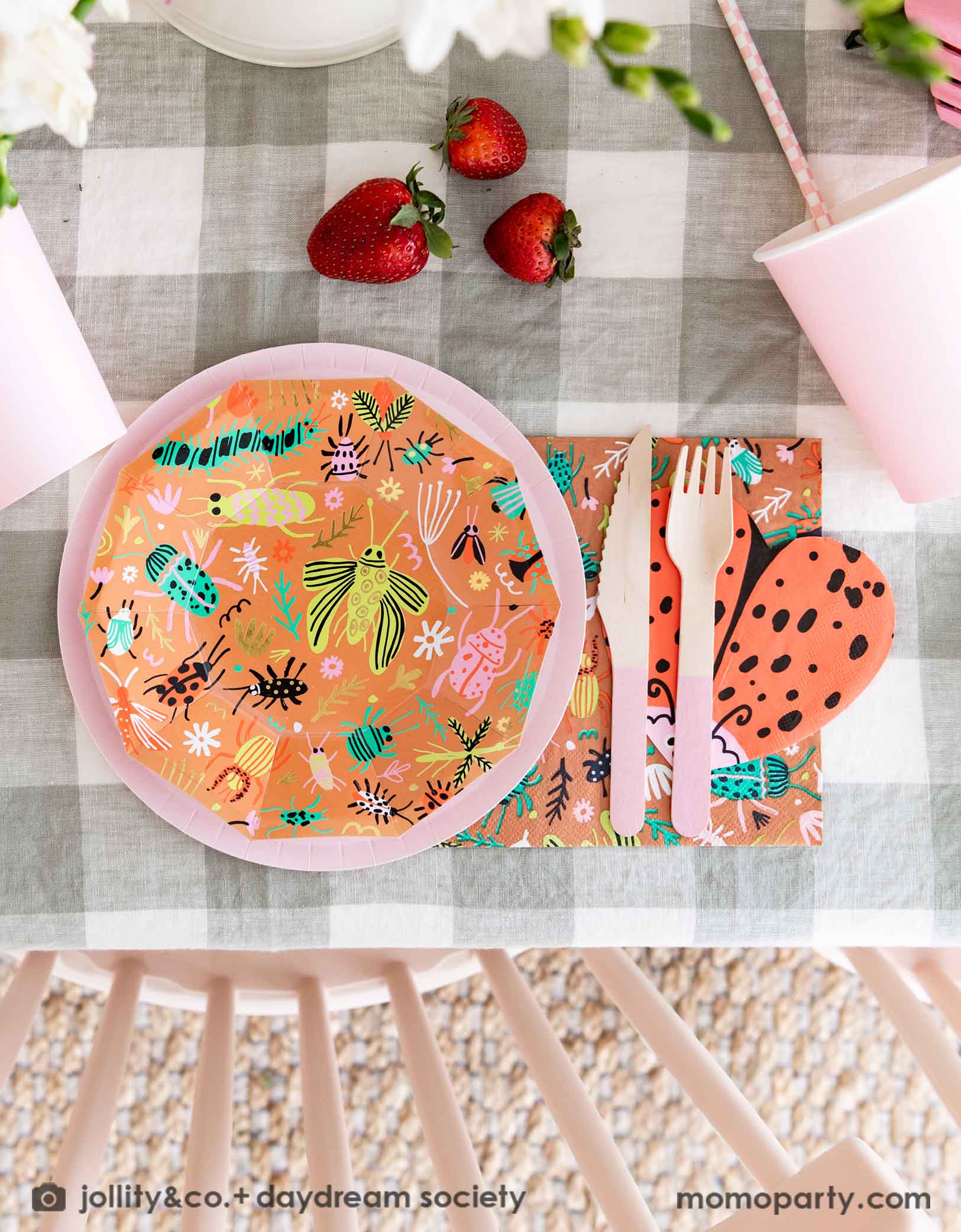 A close up shot of a cute kid's bug insect themed party table set up featuring Momo Party's backyard bug patterned plates, napkins paired with pink party cups, cutlery and plates. With an olive green gingham tablecloth under and a beautiful flower bouquet, this makes a perfect inspiration for a modern take of a kid's bug themed celebration.