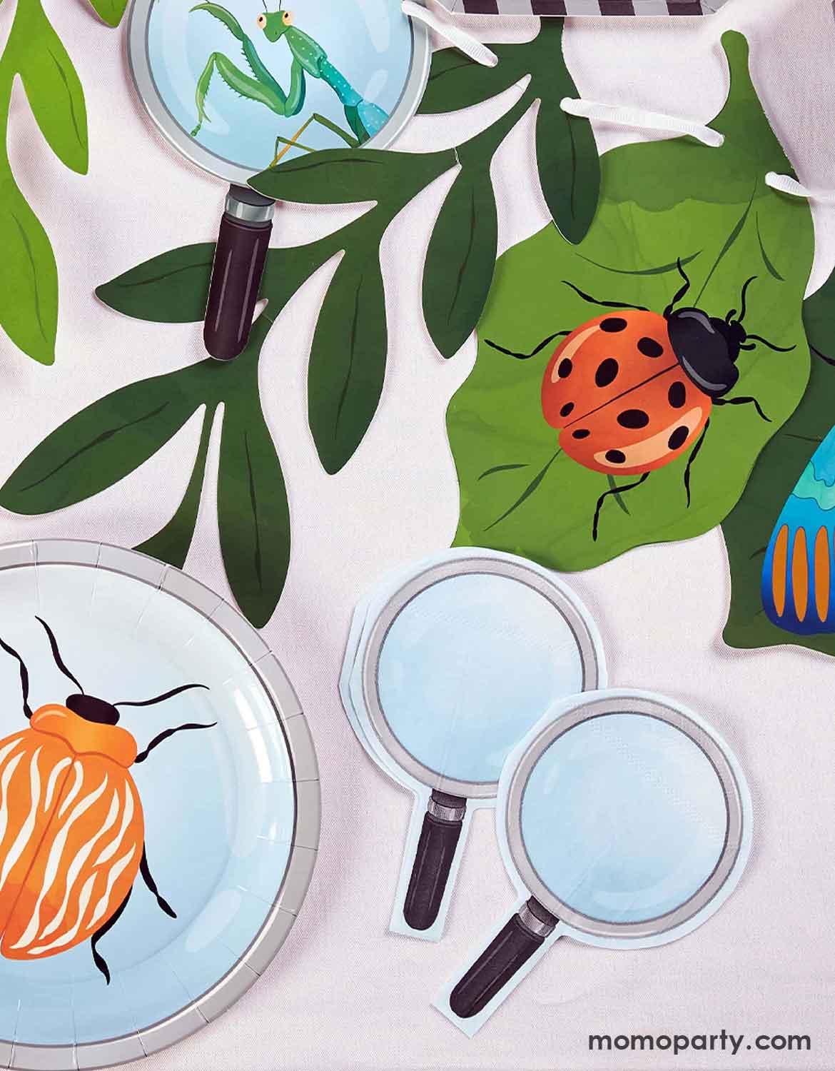 An overhead shot of multiple kid's bug themed party supplies from Momo Party laid on a white table including insect themed party banner,  magnifying glass shaped napkins, and a beetle round party plate.