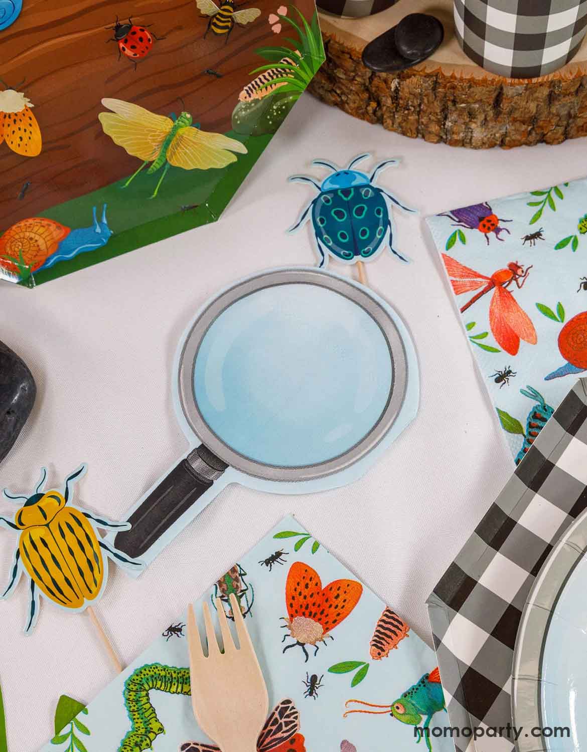 Children's tableware set with insect-themed design on a white surface. An overhead shot of multiple kid's bug themed party supplies from Momo Party laid on a white table including insect themed party plates,  magnifying glass shaped napkins, and insect themed cupcake toppers.