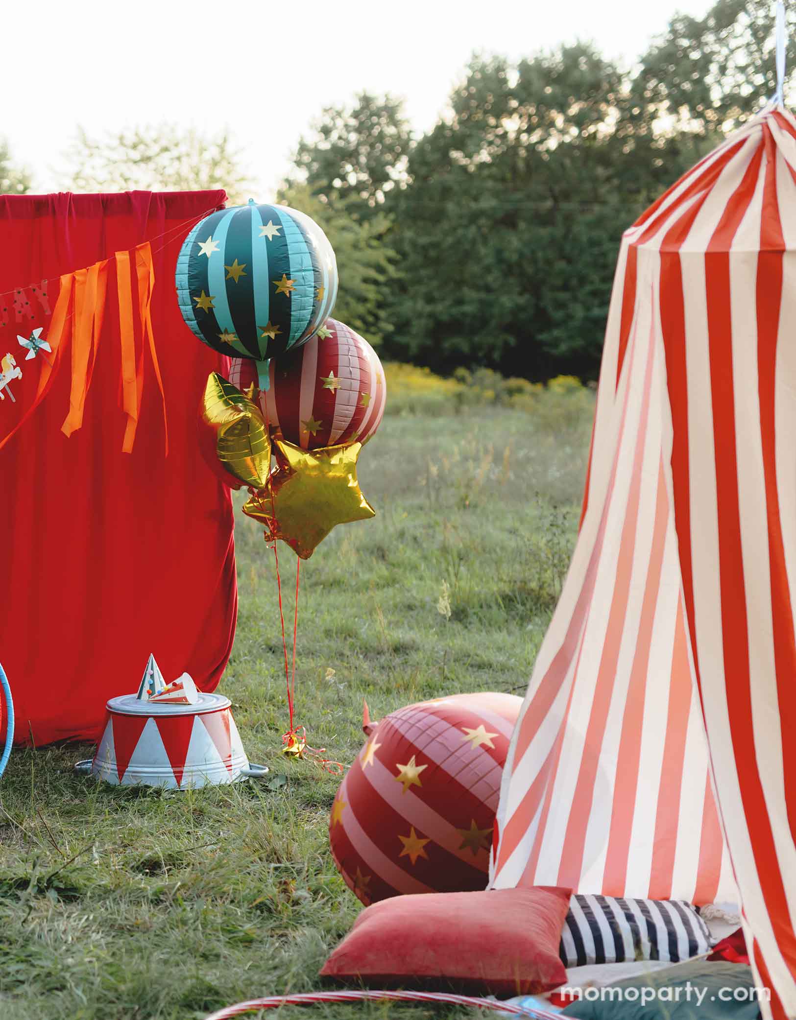 A vintage circus themed party set up in the park featuring various carnival circus themed party decorations from Momo Party including the red striped globe foil balloon, the green striped globe foil balloon and gold star foil balloons floating next to the red curtain party backdrop. In the front there's a red and white striped circus themed play tent for kids with some colorful balloons and a red striped globe foil balloon next to it. Making this a whimsical and playful setting for a fun kid's circus party.