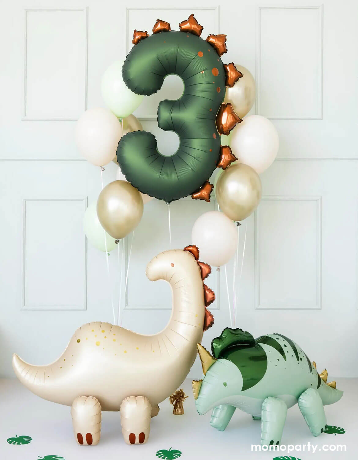 An adorable dinosaur-themed balloon decoration featuring Momo Party's olive green number three dinosaur balloon along with color-coordinated latex balloons floating against a white wall. On the floor, there are two standing dinosaur shaped foil balloons from Momo Party with palm leaf cutouts around, making this a cute set up for kid's dinosaur themed third birthday celebrations.  