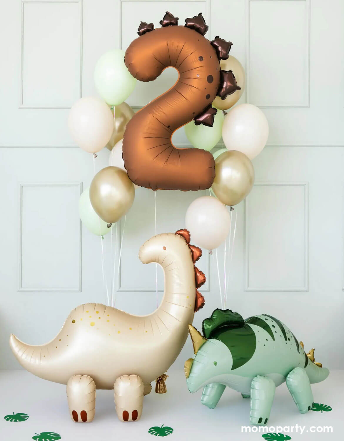 An adorable dinosaur-themed balloon decoration featuring Momo Party's matte brown number two dinosaur balloon along with color-coordinated latex balloons floating against a white wall. On the floor, there are two standing dinosaur shaped foil balloons from Momo Party with palm leaf cutouts around, making this a cute set up for toddlers dinosaur themed second birthday celebrations.  