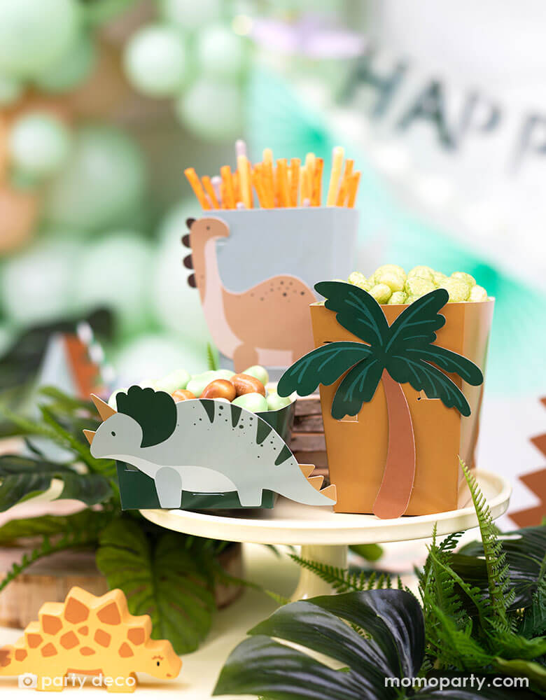 A kid's dinosaur themed party table features Momo Party's dinosaur snack boxes with cute dinosaur and palm tree designs in soft colors on the side. Around the table are some tropical plants decorated, creating a pre historical vibe. In the back the are some mint green party balloons and Happy Birthday banner.