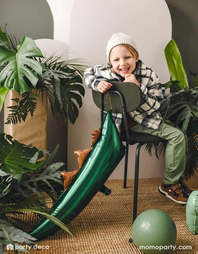 A boy dressed in green outfit sitting on a green metal chair decorated with Momo Party's 29" x 10" dark green dinosaur tail shaped foil balloon. The room is dressed with lots of tropical plants and sage green party balloons, setting a great prehistoric scene for a kid's dinosaur themed party celebration!