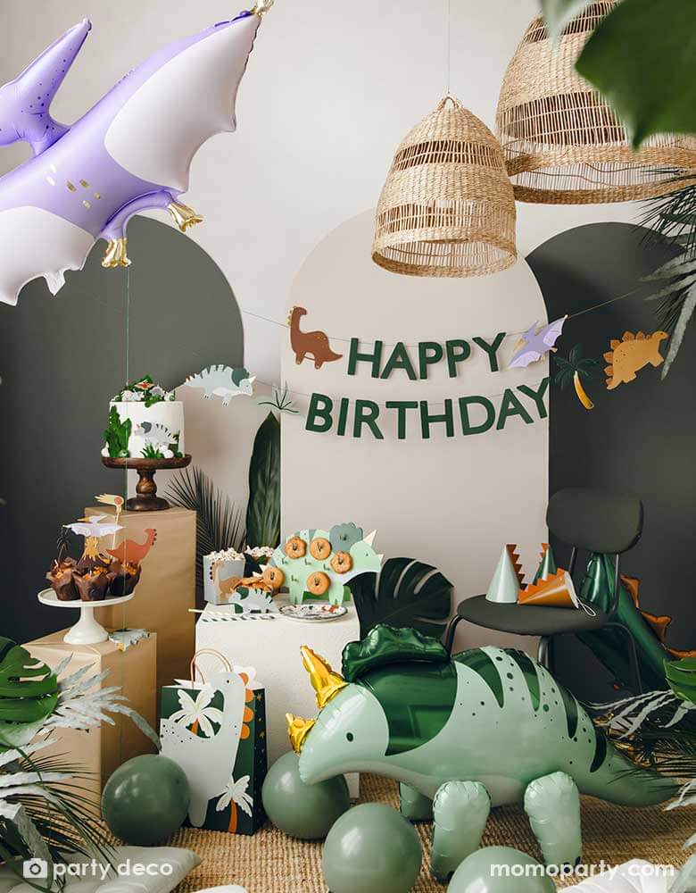 An adorable kid's dinosaur themed party set up featuring Momo Party's dinosaur themed party supplies, decorations and balloons by Party Deco including a mint Triceratops shaped foil balloon, a lilac Pterodactyl shaped foil balloon, sage green party balloons, various dinosaur cake toppers, dinosaur spikes party hats, and a dinosaur happy birthday banner hung on the party backdrop wall. All makes a cute and modern inspo for kid's dinosaur themed birthday party.