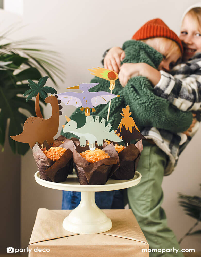 Two boys celebrating and holding each other each other at a dinosaur themed birthday party, in front of them are a few muffins topped with Momo Party's dinosaur themed cupcake toppers featuring dinosaurs, a volcano and a palm tree, setting a great prehistorical theme to a kid's dinosaur birthday party.