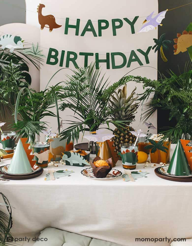 An adorable kid's dinosaur themed party set up featuring Momo Party's dinosaur themed party supplies and decorations by Party Deco including dinosaur paper plates, cup sleeves, dinosaur cake toppers, dinosaur spikes party hats, dinosaur napkins and a dinosaur happy birthday banner hung on the party backdrop wall. With a lot of tropical plants as the centerpiece, it makes a cute and modern inspo for kid's dinosaur themed birthday party.
