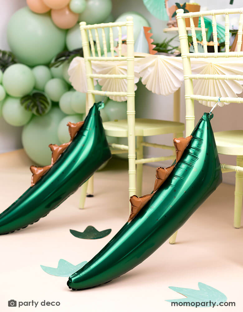 A kid's dinosaur themed birthday party table featuring party chairs decorated with Momo Party's 29" x 10" dark green dinosaur tail shaped foil balloon. On the floor there are dinosaur footprint stickers to set a prehistoric scene for a kid's dinosaur themed bash. With mint and blush balloon garland in the back this makes a perfect inspiration for a THREE-REX third birthday party bash! 