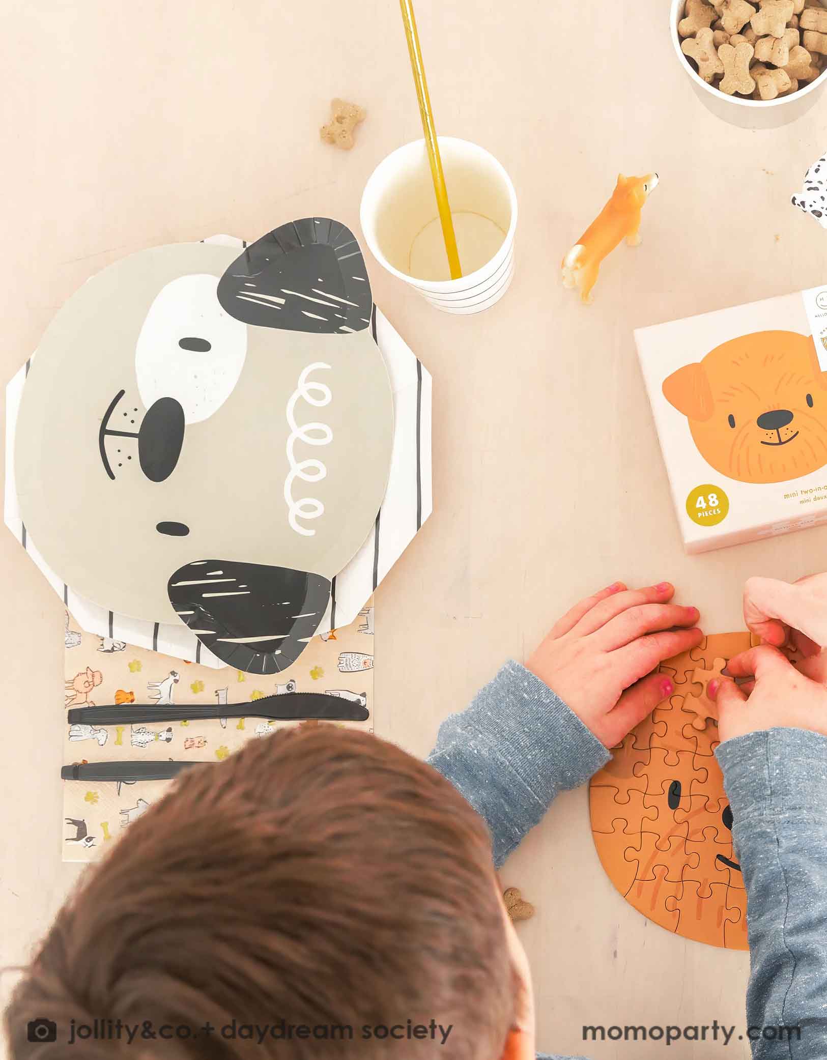 A little boy playing a puppy head shaped puzzle on a beige dining table featuring Momo Party's cute dogs head shaped paper plate paired with a black striped paper plate, along with a napkin in modern illustrations with a warm neutral color palette of dog illustrations, with black utensil set. These modern Dog Birthday Party Supplies are perfect for a dog lover, a dog themed birthday party, Let's Pawty birthday party, adorable puppy themed party.