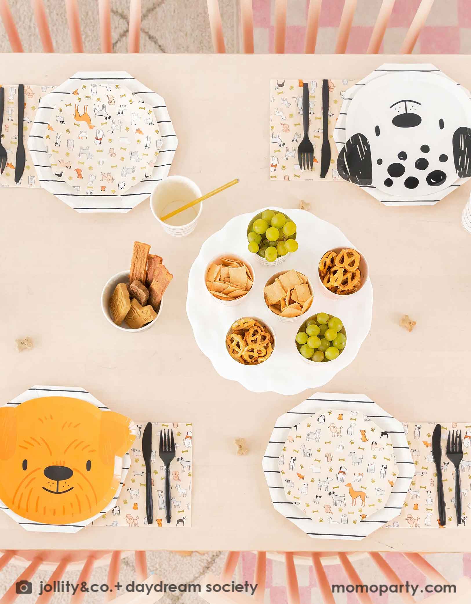 An overhead shot of Momo Party's Bow Wow Dogs themed party tablewares featuring the puppy head shaped Paper plates and Bow Wow dog small plates with black striped paper plates and cups with gold foil straw. With black utensils and some kid's snacks including grapes, pretzels, chips on the table, these warm neutral color themed modern Dog Birthday Party Supplies are perfect for a dog lover, a dog themed birthday party for kids.