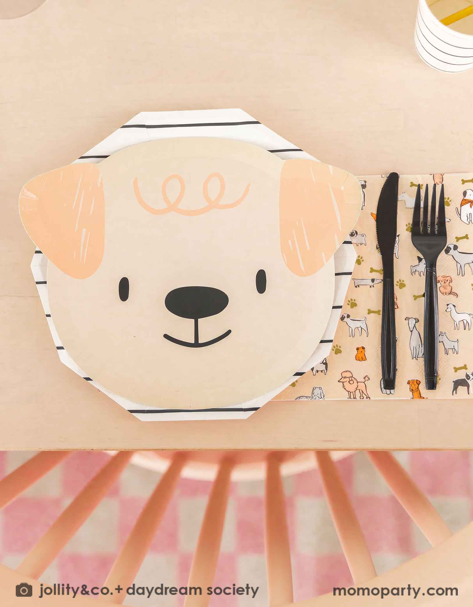 A beige dining table featuring Momo Party's cute dogs head shaped paper plate paired with a black striped paper plate, along with a napkin in modern illustrations with a warm neutral color palette of dog illustrations, with black utensil set. These modern Dog Birthday Party Supplies are perfect for a dog lover, a dog themed birthday party, Let's Pawty birthday party, adorable puppy themed party.