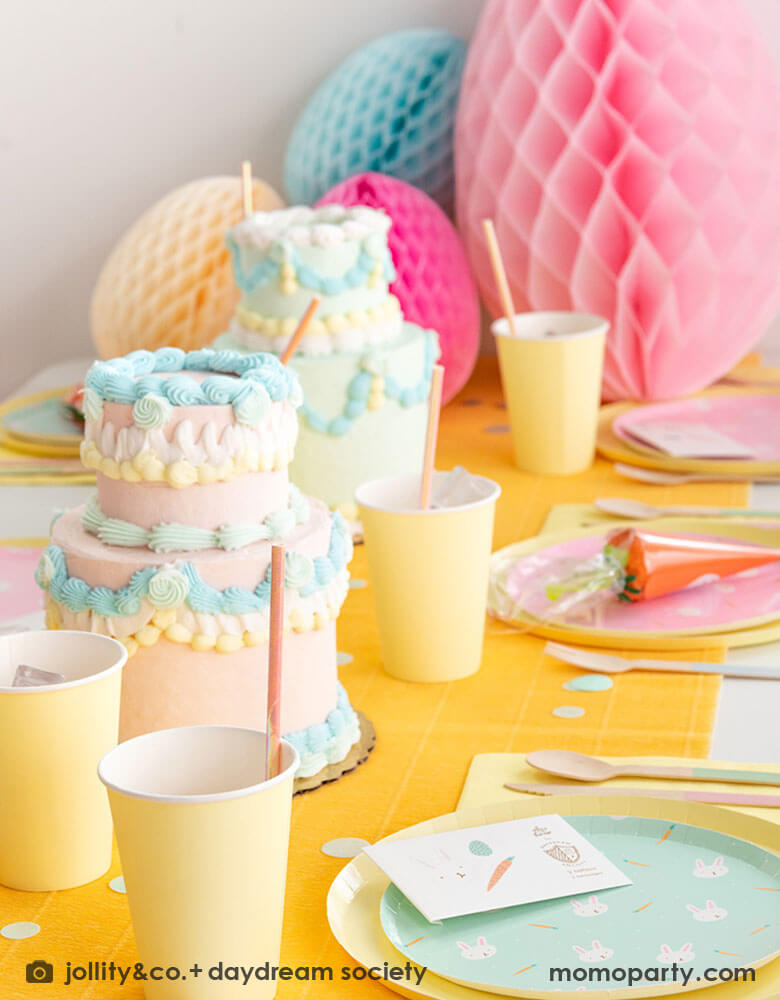 A kid's Easter party table featuring Momo Party's Easter Fun party collection by Daydream Society including Easter Fun plates in mint and pink, Easter Fun temporary tattoos, pale yellow dinner plates and party cups. On the table there are two 2-tier vintage inspired buttercream cakes with pastel piping. In the back there are multiple giant honeycomb Easter egg decorations in pastel colors of pink, ivory, and pale blue, making this a perfect inspiration for Easter brunch hosting occasion!