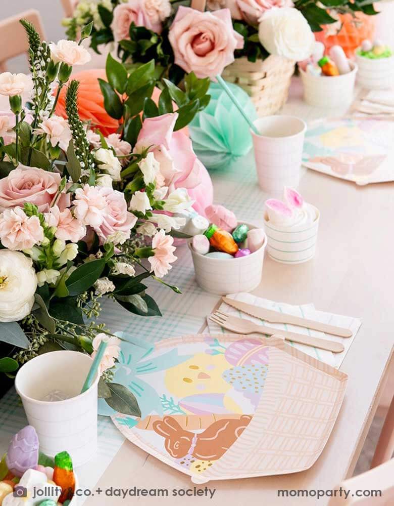 An adorable kid's friendly Easter table setting features Momo Party's Easter Fun basket shaped plates, mint striped plates, party cups by Daydream Society. In the middle there's light blue gingham table runner, Easter Egg tissue honeycomb in mint and peach and a beautiful Easter flower arrangement in a basket. On the table there's also small treat cups filled with Easter candies and chocolates, making this an adorable Easter tablescape.