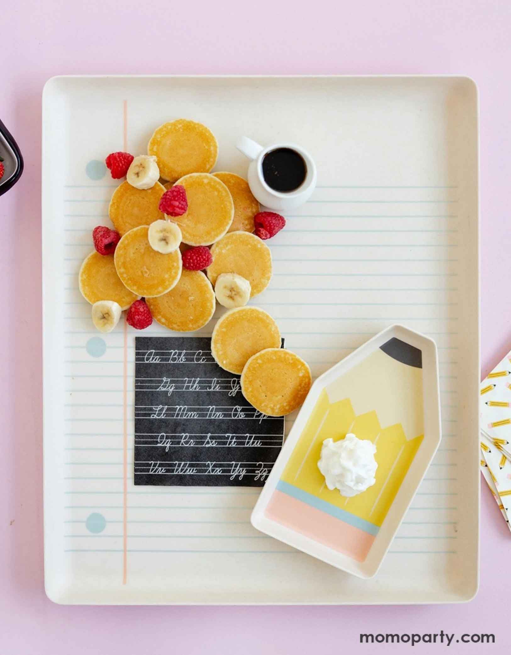 An overhead shot of Momo Party's 17" x 14" Paper and Pencil Bamboo Tray Set filled with a bunch of petite pancakes, banana bites, raspberries and a small syrup pourer. On the notebook bamboo tray is the black Cursive Abcs Paper Small Napkin and the pencil shaped bamboo try with a dollop of whip cream. This makes a great inspiration idea for a special first day of school breakfast for kids to celebrate the new school year!
