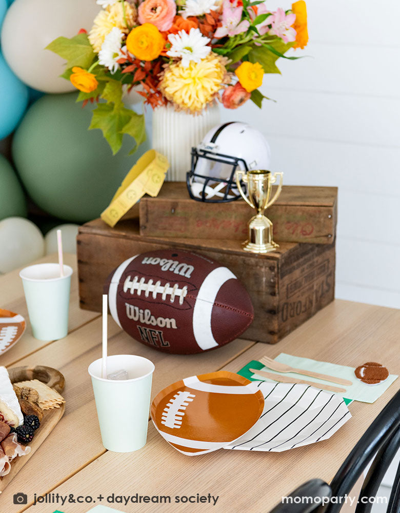 A modern kid's football birthday party table filled with Momo Party's football themed party tableware by Daydream Society including the die-cut football shaped plates, black striped plates, green and mint napkins and party cups. In the center of the table there's a cheese charcuterie board and some decorations like a football, a trophy and a football helmet as the centerpiece. On the wall there are some old school football posters and color matching balloons to add some festivities to the celebration!