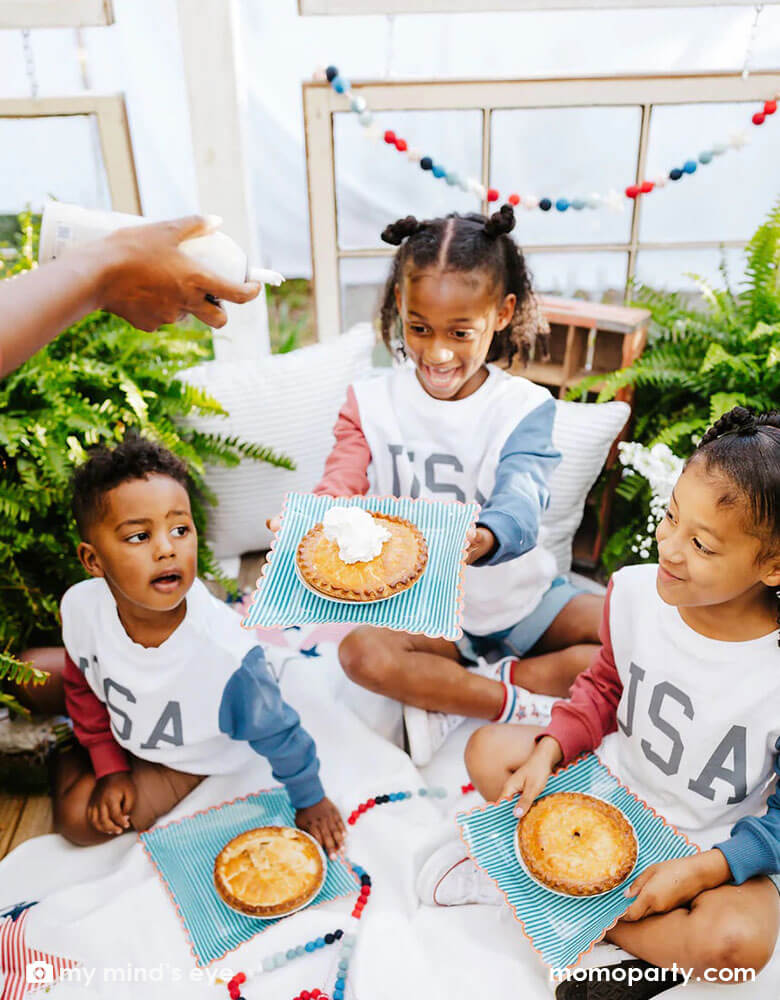 Three siblings in their USA sweatshirts holding Momo Party's red with blue stripe plates accented by a red scalloped edge. Designed by My Mind's Eye, these plates are in a perfect size for summer treats, including hand pies, hot dogs, burgers and more for a 4th of July cookout!