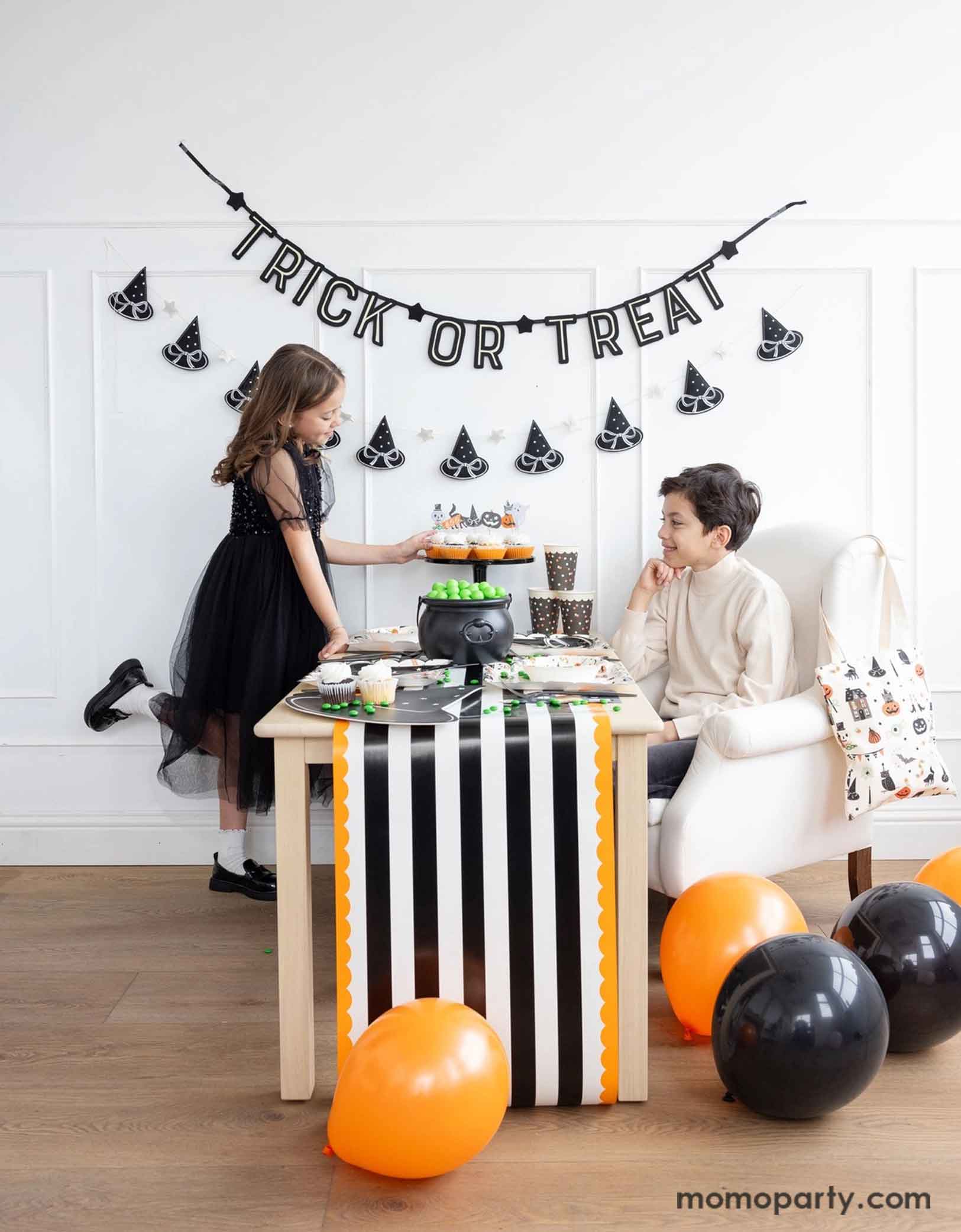 Two children at a Halloween-themed party with Momo Party's 'Trick or Treat' banner and the witch's hat garland hung on the white wall. There's a table decorated with multiple Halloween party supplies by Momo Party including witch's hat shaped bamboo tray, party cups and orange, black and white striped table runner draping from the table which some orange and black balloons next to. A cute and simple set up for kids to celebration Halloween at home.