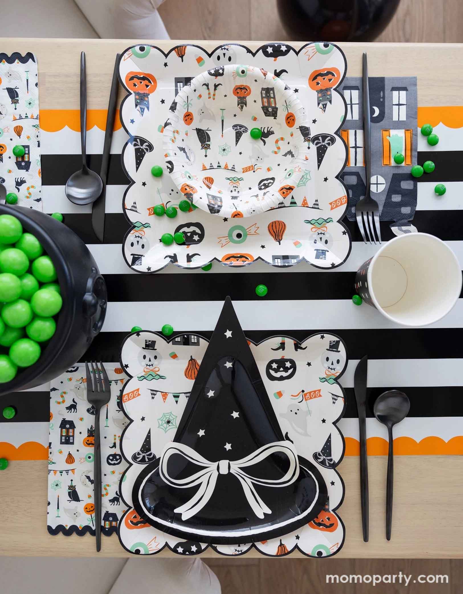 A overhead shot of a festive Halloween-themed table setting with the witch hat shaped plate, Trick or Treat dinner plates and bowls, the haunted mansion shaped napkins and black cutlery set on a black orange and white striped table runner fro Momo Party. On the table there's a cauldron filled with green gum balls and some green candies spread across the table. A perfect set up for kid's friendly Halloween celebration.