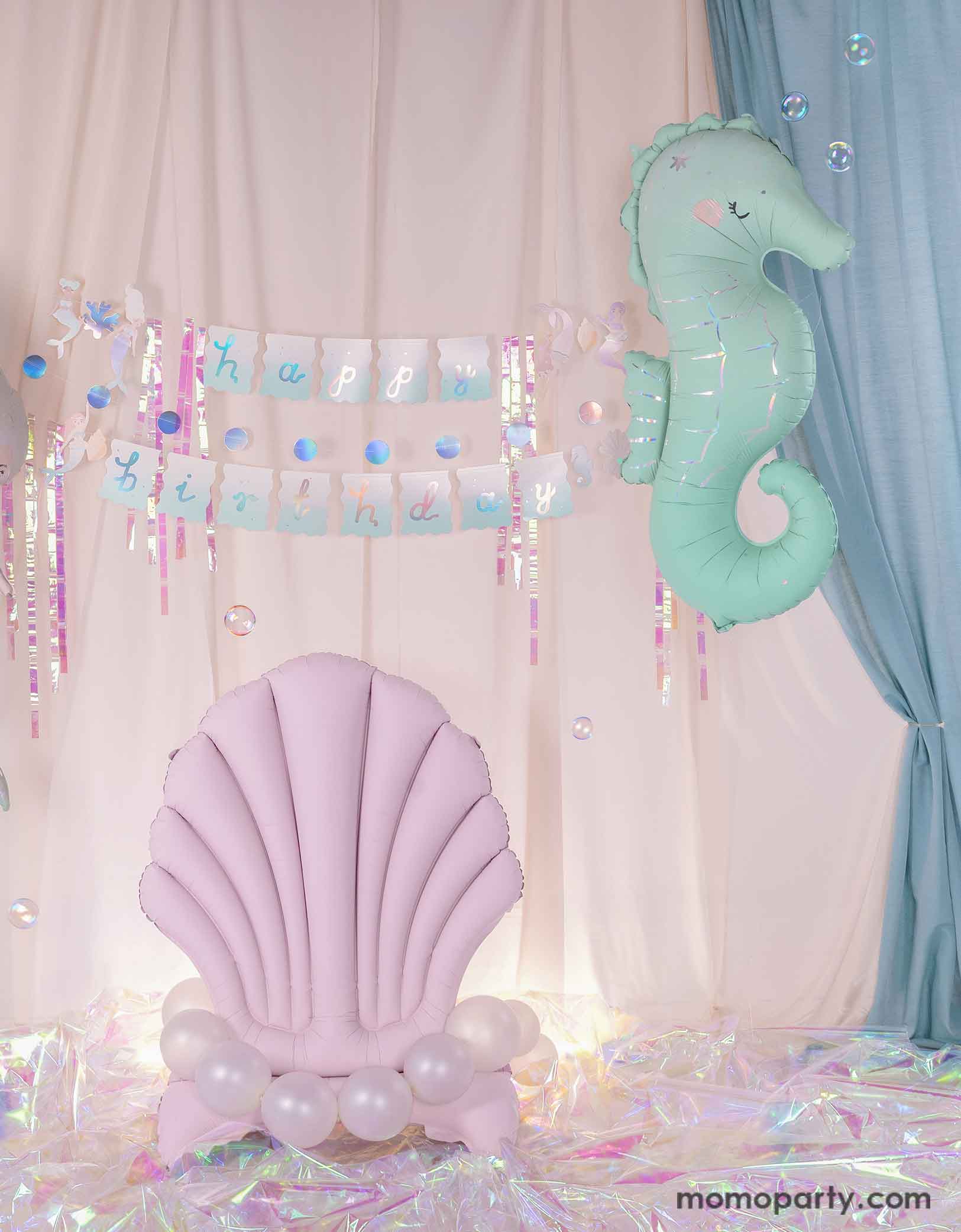 An enchanting and dreamy kid's mermaid party decoration set up featuring Momo Party's under the sea mermaid themed party supplies including a floating mint seahorse balloon, the lilac seashell standing foil balloon adorn with pearl white balloons and the mermaid happy birthday part garland set with iridescent streamers. With a white and light blue curtain in the back as the backdrop and iridescent cellophane on the floor, making this a magical setting for kid's mermaid party.