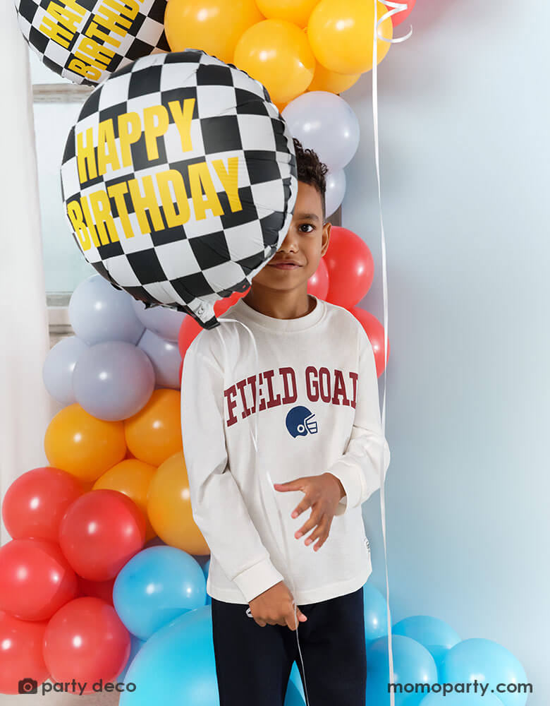 A boy holding Momo Party's 14" checkered flag foil balloon with gold foil Happy Birthday written on it. He is standing in front of a festive balloon garland in race car inspired colors, ready for a fun race car birthday bash to begin!