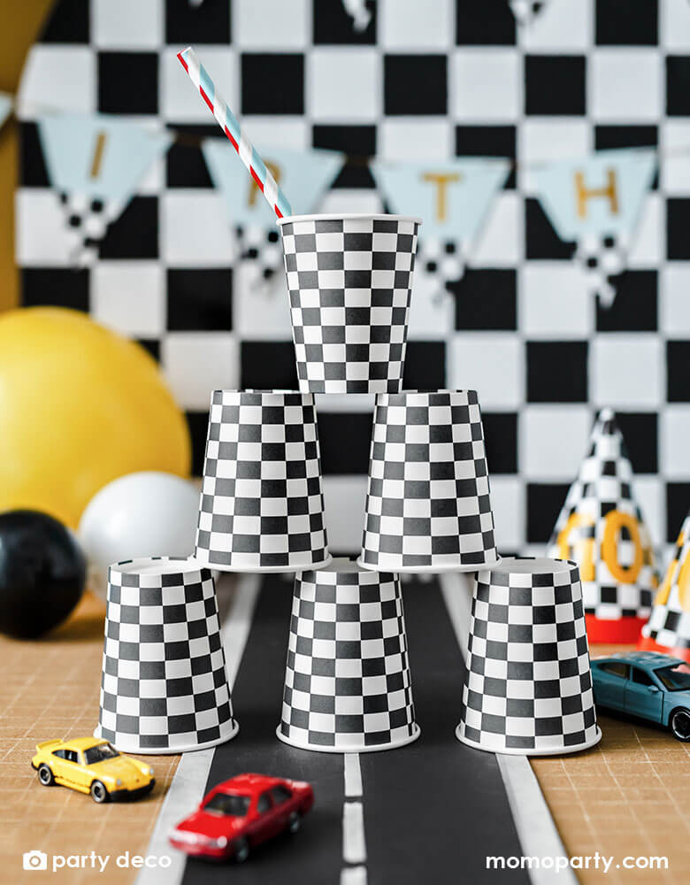 A kid's race car party featuring 6 race car checkered flag party cups stacked in three rows. Around the cups there are some toy cars and some checkered party hats. In the back there are some bright colored birthday balloons and checkered backdrop with a happy birthday banner hung on it.