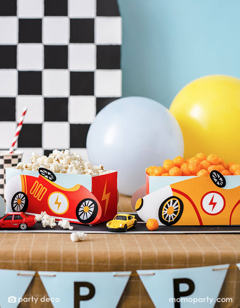 A modern kid's race car themed party table features Momo Party's race car snack boxes in yellow and red, filled with popcorn and cheese balls. On the table there are checkered pattern party cups and race track table runner, with toy cars decorated. In the back there's a checkered backdrop decorated with some party balloons in light blue and yellow, making this a perfect inspo for kid's race car themed parties including a "Two Fast" second birthday party or a "Fast One" first birthday party."