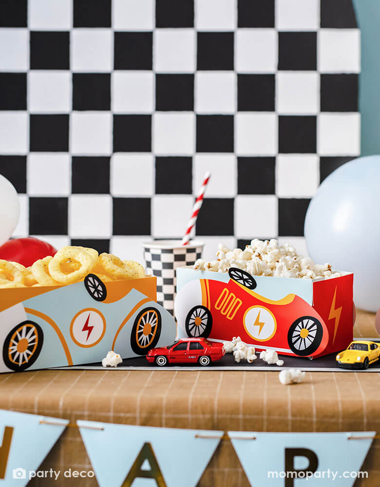 A modern kid's race car themed party table features Momo Party's race car snack boxes in light blue and red, filled with popcorn and onion ring chips. On the table there are checkered pattern party cups and race track table runner, with toy cars decorated. In the back there's a checkered backdrop decorated with some party balloons, making this a perfect inspo for kid's race car themed parties including a "Two Fast" second birthday party or a "Fast One" first birthday party."