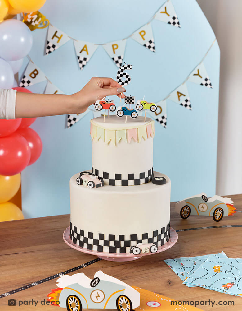 A race car themed birthday cake decorated with checkered pattern at the bottom topped with Momo Party's race car candles, a hand putting a checkered number 6 candle on the cake. In the back there's a race car themed trophy happy birthday banner hung on the wall. On the table there are some race car themed paper napkins and race car shaped party invitation. Makes it a great inspiration for kid's race car birthday party.