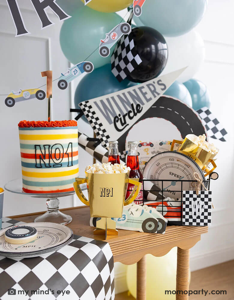 A festive corner of a race car party table filled with race car themed party supplies from Momo Party including speedometer shaped plates, race car shaped napkins, race car pattern plates, trophy shaped treat boxes with popcorn, and the "Winner's Circle"felt party pennant. Next to the party supplies is a colorful yet modern buttercream cake which has "No. 1" written on it. In the back is a colorful balloon garland in blue, black, yellow colors adorned with a race car party banner and some checkered flags.