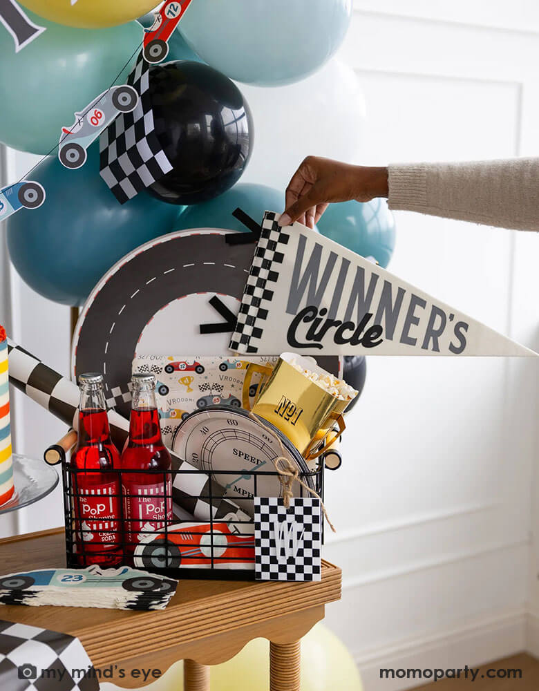 An occasion bin filled with race car themed party supplies from Momo Party including race track placemats, speedometer shaped paper plates, checkered table runner, race car pattern square plates, race car shaped napkins, and trophy shaped treat boxes. A hand is reaching to grab the felt "Winner's Circle" party pennant from the basket. Around the basket is a colorful buttercream cake and some party balloons and checkered flags decorated behind the table - a perfect inspo for kid's race car themed parties.
