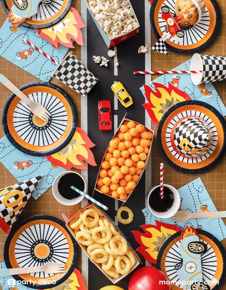 A kid's race car themed party table filled with race car themed party supplies from Momo Party including the wheel shaped plates, pastel blue race car large napkin, and checkered party cups. On the table there are muffins topped with race car themed cupcake toppers and a red vintage race car shaped snack holder filled with popcorn. All laid on a brown checkered tablecloth adorned with race car track in the middle with some party snacks and toy race cars, making these a perfect inspo for a car themed party!