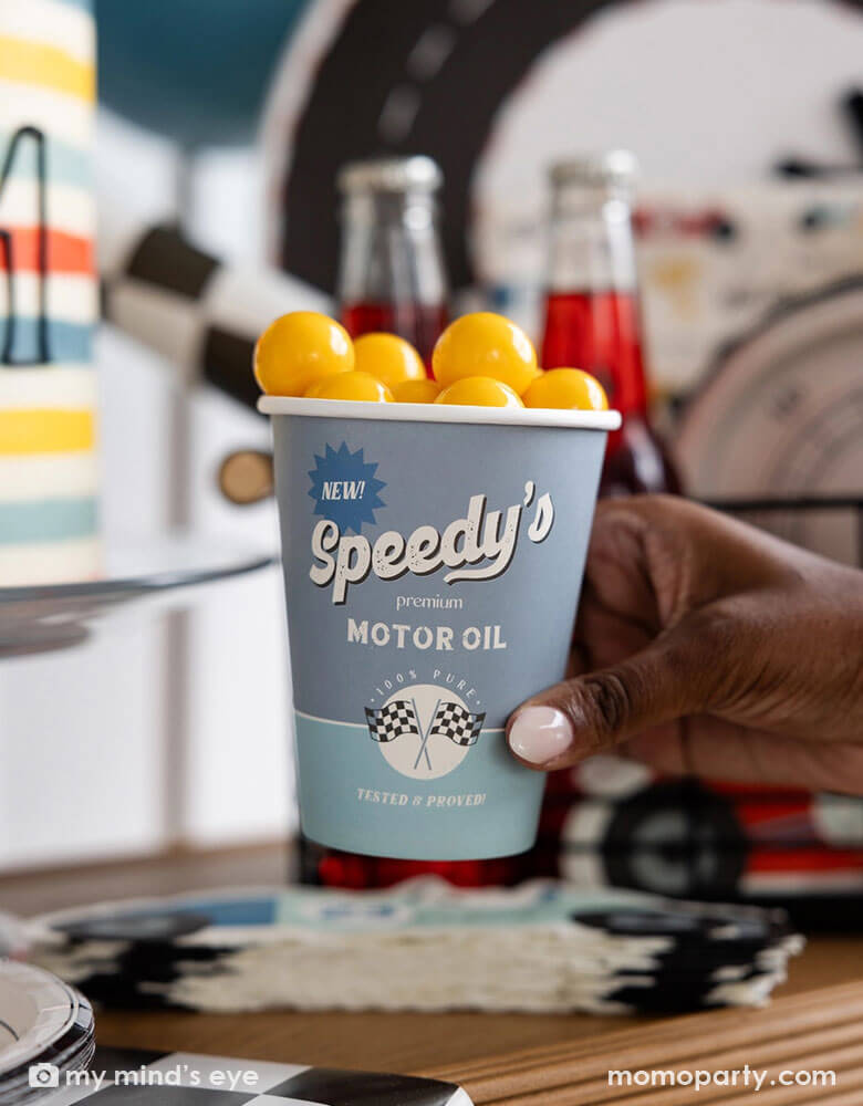 A kid's race car party featuring Momo Party's 12 oz race car cup by My Mind's Eye. Comes in a set of 8 paper cups in light blue and mint with Speedy's motor oil logo on it and some checkered flags design. In the car there are yellow gum balls as kid's treats and in the back you can see some festive race car themed party decorations, birthday cakes, treats and drinks.