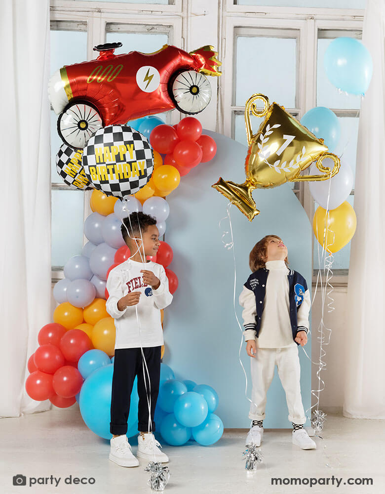 Two boys holding Momo Party's race car themed foil balloons including a red vintage race car shaped balloon, two checkered flag happy birthday foil balloons and a gold trophy shaped foil balloon, both kids standing in front of a pastel blue party backdrop wall which is decorated with colorful balloon garland with race car inspired colors including red, light blue, orange, gray and yellow. All makes this a festive and fun kid's birthday celebration.
