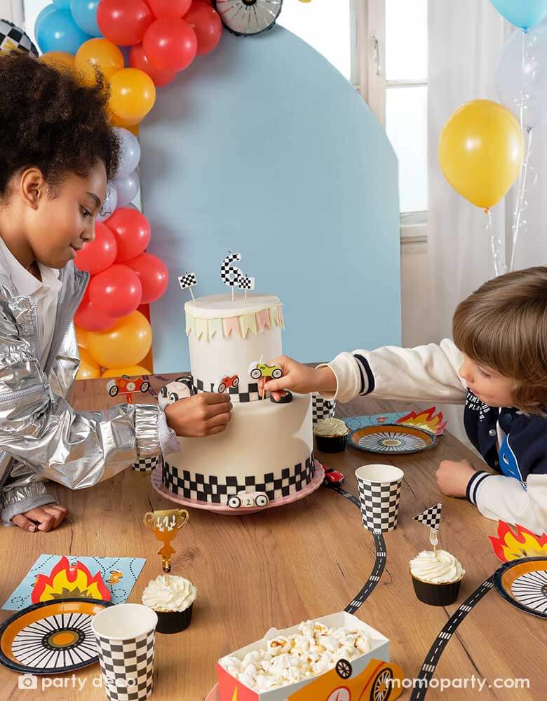 Two kids putting race car shaped candles on a race car themed birthday cake, around the cake there are lots of race car themed party supplies including Momo Party's wheel shaped plates, napkins, checkered party cups, cupcakes topped with race car themed toppers, and race car shaped snack holders. In the back there's a pastel blue backdrop adorned with colorful balloon garland in race car themed colors.
