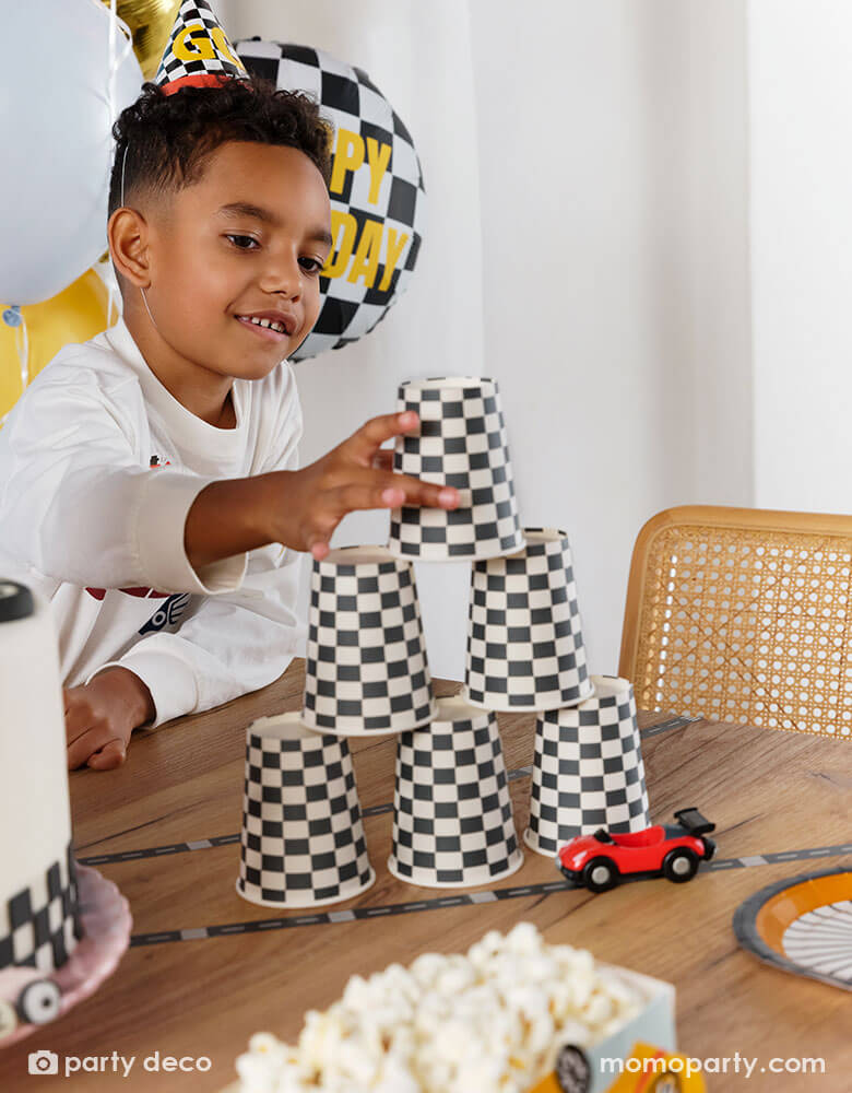 A boy wearing Momo Party's checkered race car party hat stacking the checkered flag party cups to make a cup tower on a party table, next to the cups are some party supplies from Momo Party including wheel shaped plates, race car track tape, and some toy cars.