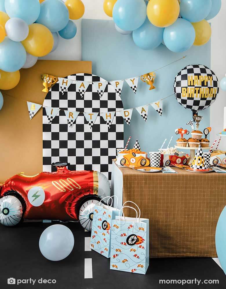 A kid's race car themed party featuring a table filled with race car themed party supplies from Momo Party including wheel shaped plates, checkered flag GO! party hats, blue race car large napkin, and checkered party cups. Behind the table is a checkered party backdrop adorned with trophy happy birthday banner with colorful balloon garland. On the floor there's a vintage red race car foil balloon setting the scene for this festive celebration. A perfect inspo for kid's Two Fast second birthday party.
