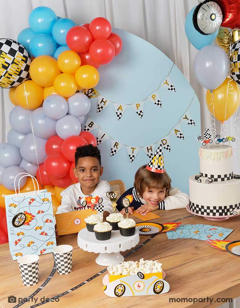 Two boys in a race car themed birthday party with them sitting at a table filled with race car themed party supplies from Momo Party including wheel shaped party plates, checkered party cups, race car themed napkins and party bags. With birthday cake and cupcakes topped with race car candles and cupcake toppers. Behind them is a blue backdrop wall decorated with trophy happy birthday banner from Momo Party and colorful balloon garland in the race car themed colors.